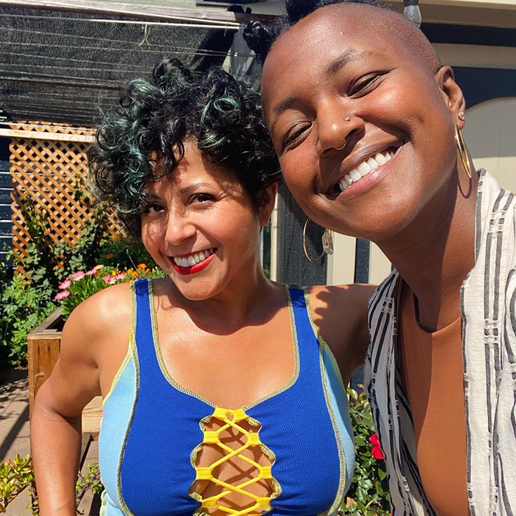 Favianna has short curly hair with blue green highlights. She is wearing a two tone blue tanktop. Favianna has her arm around her close friend Jamila who is also smiling as the sun hits their faces.